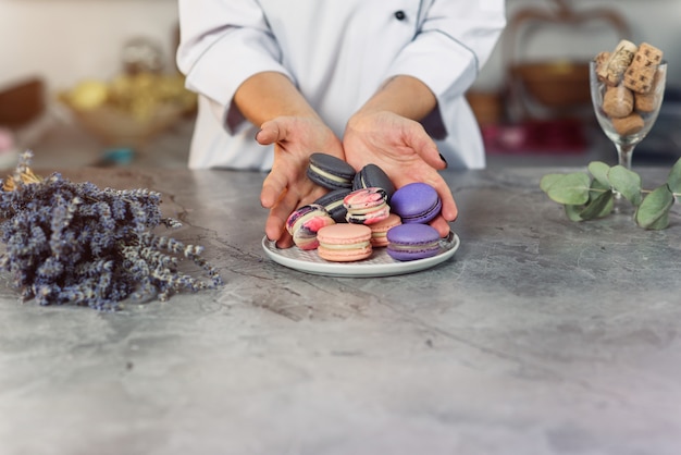 Le mani femminili di Baker versano macarons colorati in un piatto bianco su un tavolo di marmo.