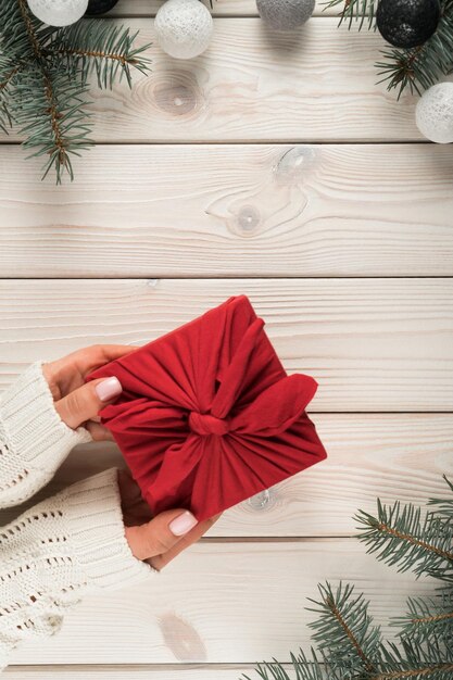 Le mani femminili in maglione bianco tengono il regalo di Natale nella vista dall'alto del pacchetto furoshiki Cornice di rami di abete e palle di albero di Natale Concetto di vacanza di Natale o Capodanno arredamento ecologico zero rifiuti