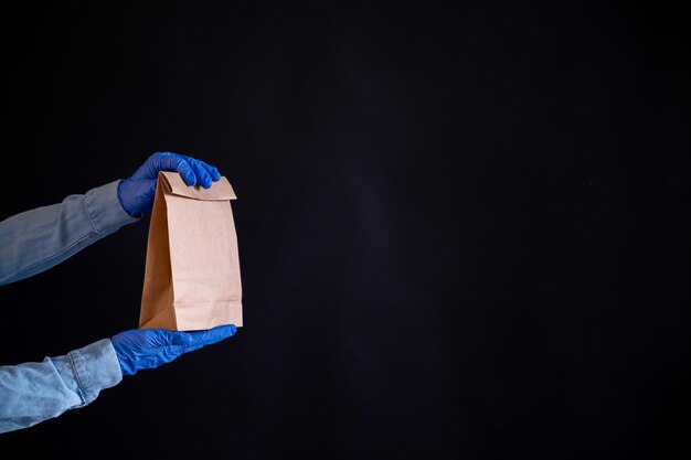 Le mani femminili in un guanto blu tengono un sacchetto di carta marrone su sfondo nero Consegna sicura di cibo a casa tua Un corriere in una camicia di jeans porge un sacchetto di cartone artigianale a un cliente