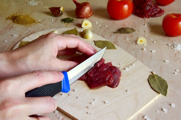 le mani femminili tagliano la carne di manzo con un coltello su un tagliere di legno nella cucina di casa.