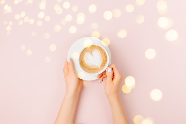 Le mani femminili tengono l'arte del cappuccino su sfondo rosa pallido