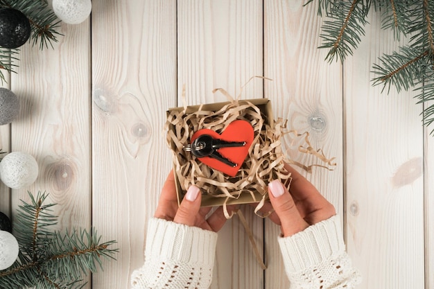 Le mani femminili tengono la scatola regalo con le chiavi di casa e cuore di carta rossa cartolina vista dall'alto Cornice da rami di abete rosso e palle di albero di Natale Acquisto di nuovi immobili Vacanze di Natale o Capodanno