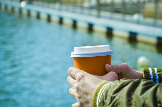 Le mani femminili tengono la tazza di cartone con caffè sullo sfondo del molo sul mare. Relax in riva al mare, passeggiate lungo la costa, caffè da asporto. Spazio per il testo. Messa a fuoco selettiva sulle mani e sulla tazza di caffè