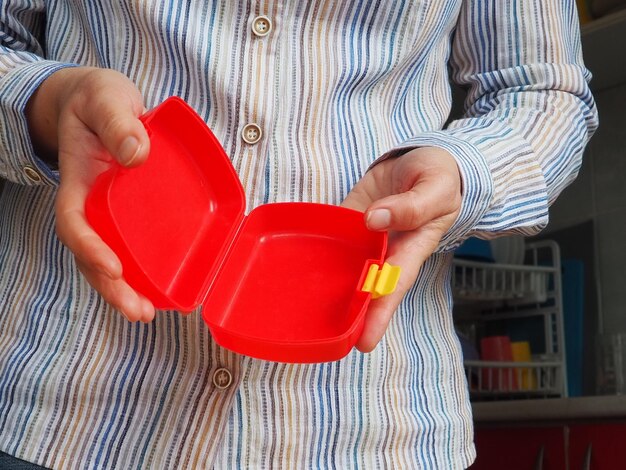 Le mani femminili tengono un contenitore o un imballaggio per il pranzo scolastico di plastica aperto rosso Utensili da cucina Una donna con una camicia a righe bianche mostra la scatola del pranzo Igiene alimentare e tema del riciclaggio