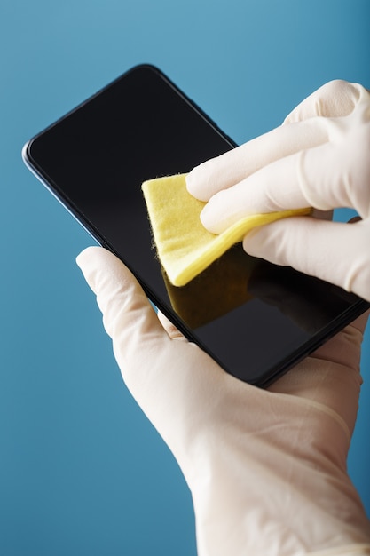 Le mani in guanti protettivi puliscono e disinfettano uno smartphone con un tovagliolo giallo su una superficie blu