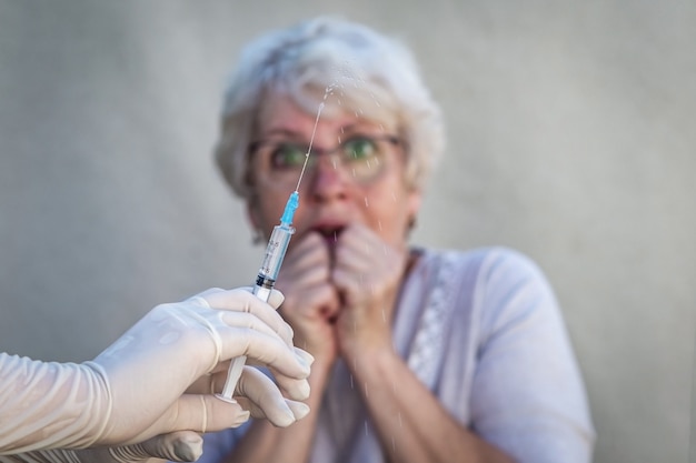 Le mani nei guanti medici tengono in mano una siringa, si preparano per la vaccinazione, sullo sfondo una donna anziana con gli occhiali ha paura di fare un'iniezione.