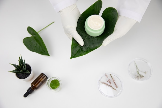 Le mani nei guanti tengono il barattolo di crema cosmetica su una foglia verde su sfondo bianco da laboratorio