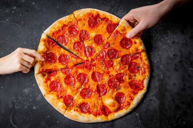 Le mani prendono i pezzi della classica pizza italiana ai peperoni su sfondo nero, vista dall'alto. Foto di alta qualità