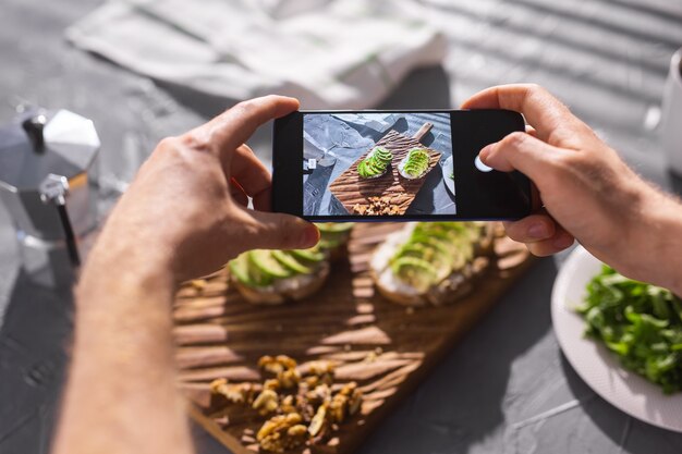 Le mani scattano foto sullo smartphone di due bei panini sani con panna acida e avocado sdraiati