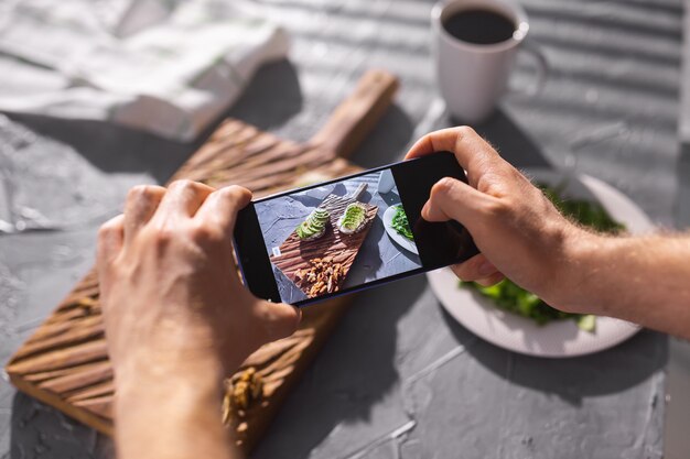 Le mani scattano foto sullo smartphone di due bei panini sani con panna acida e avocado sdraiati