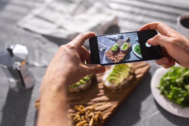 Le mani scattano foto sullo smartphone di due bellissimi panini sani con panna acida e avocado che si trovano a bordo sul tavolo. Social media e concetto di cibo