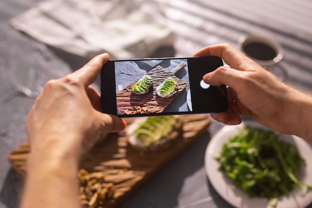 Le mani scattano foto sullo smartphone di due bellissimi panini sani con panna acida e avocado che si trovano a bordo sul tavolo. Social media e concetto di cibo