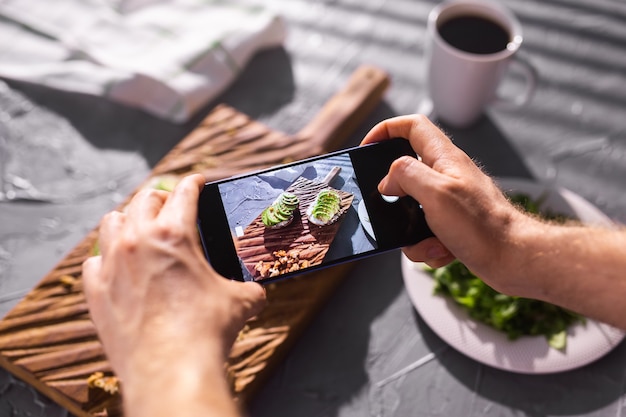 Le mani scattano foto sullo smartphone di due bellissimi panini sani con panna acida e avocado che si trovano a bordo sul tavolo. Social media e concetto di cibo