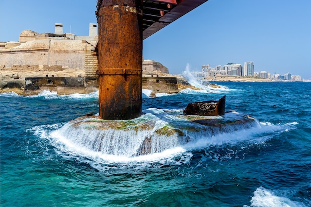 Le onde turchesi del Mar Mediterraneo si infrangono sul supporto metallico del ponte di Sant'Elmo a Malta, La Valletta.