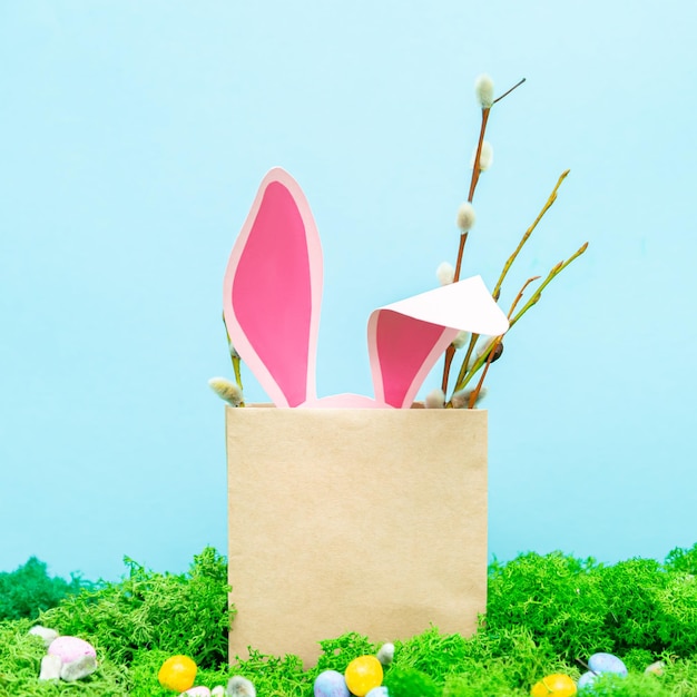 Le orecchie del coniglio rosa sporgono dalla borsa Biglietto di auguri primaverile con erba e fiori su sfondo blu Simboli pasquali