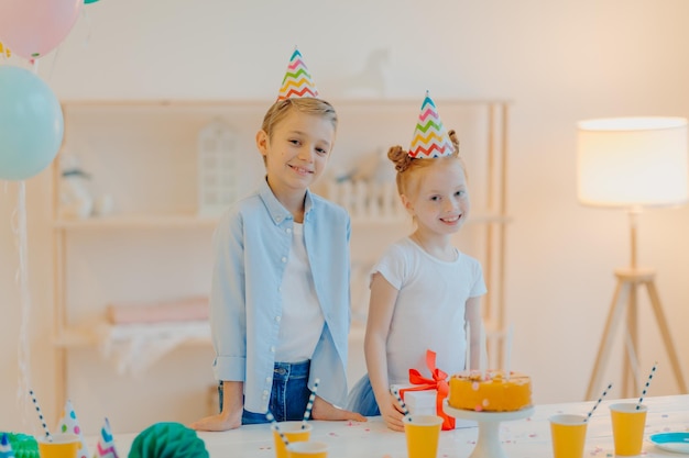 Le riprese in interni di piccole ragazze e ragazzi indossano cappelli da festa in piedi vicino a un tavolo festivo con bicchieri di carta per torte e presenti festeggiano il compleanno insieme posano in una spaziosa stanza bianca Concetto di celebrazione