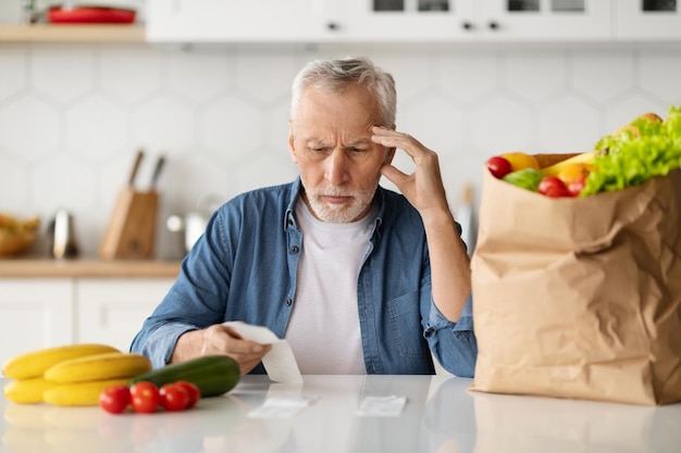 Le spese di generi alimentari hanno sconvolto l'uomo anziano in cucina che controlla il conto dopo la spesa alimentare