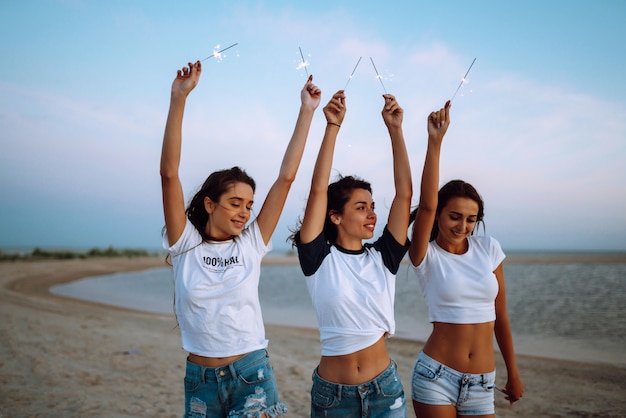 Le stelle filanti nelle mani di giovani ragazze sulla spiaggia. Tre ragazze che godono del partito sulla spiaggia con le stelle filante. Vacanze estive, vacanze, relax e lifestyle concept.