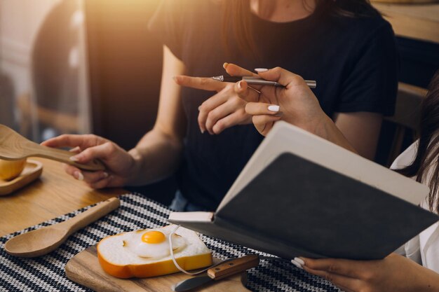 Le studentesse notano dai libri della biblioteca della ragazza asiatica seduta al divano utilizzando il computer portatile e il tablet per cercare informazioni online nel soggiorno