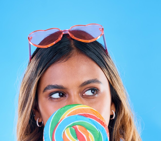 Lecca-lecca pensante e una donna su sfondo blu in studio con occhiali a cuore per la moda Idea caramelle e dolci con una giovane donna attraente che mangia uno spuntino gigante mentre si sente premurosa