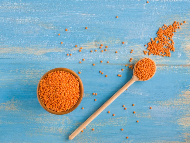 Lenticchia arancione rossa o canadese sulla ciotola di legno e cucchiaio su sfondo blu. Dieta vegetariana. Fonte di fibre per abbassare il colesterolo. Lenticchie sgusciate con un mantello di semi marrone e un cotiledone rosso-arancio.