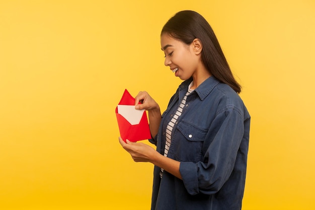 Lettera d'amore a San Valentino. Felice bella ragazza in camicia di jeans tirando la lettera dalla busta rossa, tenendo in mano il biglietto di auguri e sorridendo con gioia. tiro in studio indoor isolato su sfondo giallo