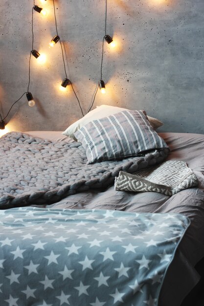 Letto con cuscini in camera soppalcata, pareti in cemento con ghirlanda di lampadine. Interni scandinavi