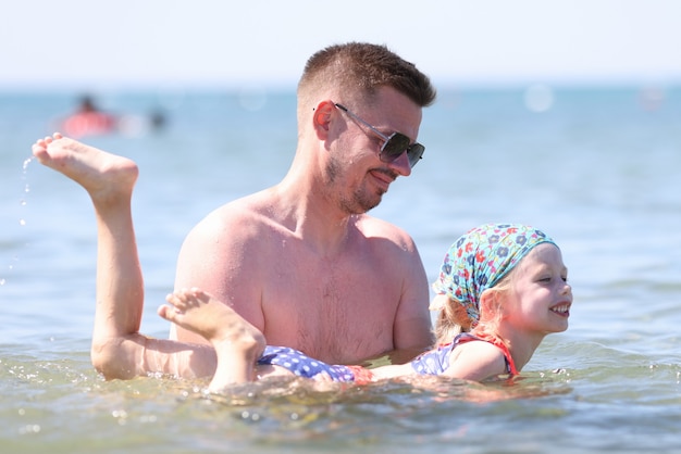 Lezione di nuoto per bambini su base individuale. L'uomo in occhiali da sole tiene la ragazza con le mani sopra l'acqua.