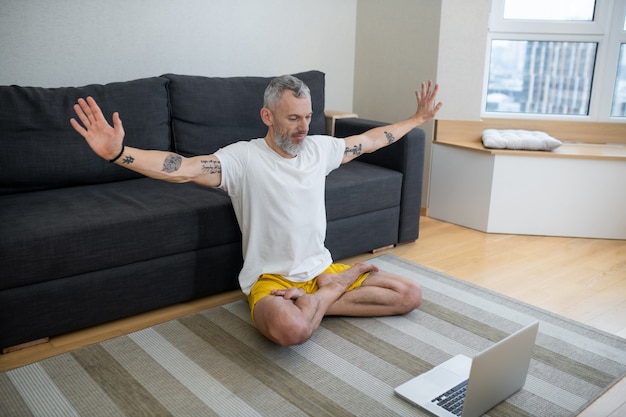 Lezioni di yoga. Un uomo maturo con una maglietta bianca seduto sul pavimento e che registra un tutorial di yoga online