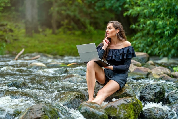 libera professionista femminile seduta su una roccia nel fiume e utilizzando un concetto di viaggio o di lavoro in vacanza con un laptop