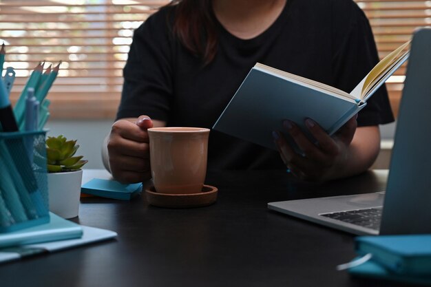 Libera professionista femminile sho ritagliata che beve caffè e legge un libro a casa.