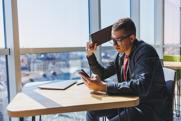 Libero professionista allegro con gli occhiali che naviga sullo smartphone e sorride mentre è seduto al tavolo con laptop e blocco note nella caffetteria durante il giorno Lavorando in remoto dall'ufficio