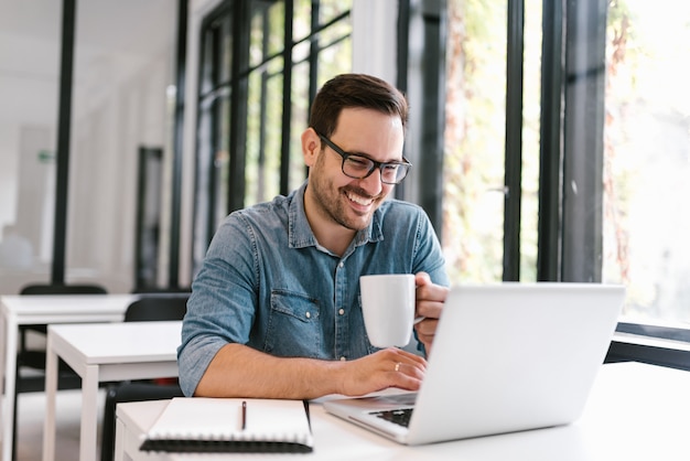 Libero professionista bello che utilizza computer portatile e che beve caffè nell&#39;ambiente moderno luminoso.