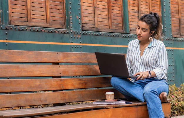 Libero professionista che lavora su un computer portatile seduto su una panchina al parco bevendo una tazza di caffè