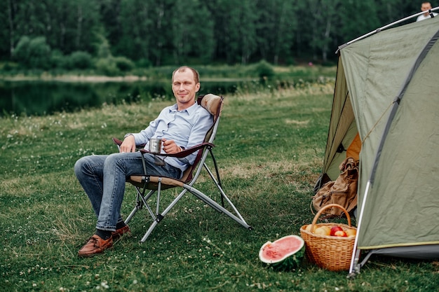 Libero professionista del giovane che si siede sulla sedia e che si rilassa davanti alla tenda al campeggio in foresta o prato. Attività all'aperto in estate. Avventura in viaggio nel parco nazionale. tempo libero, vacanza, relax