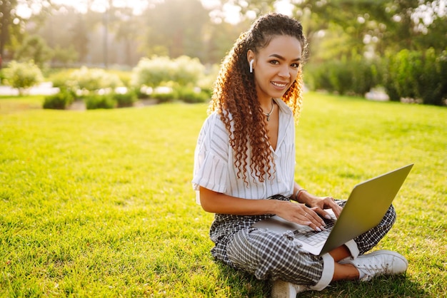 Libero professionista della giovane donna che si siede sull'erba verde con il computer portatile Istruzione online