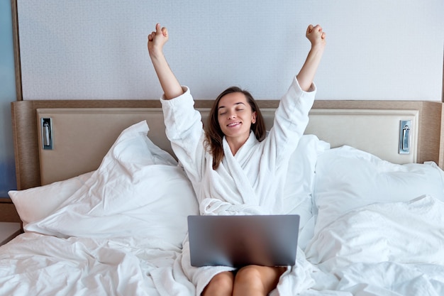 Libero professionista giovane carino calma sorridente sognando donna con le mani alzate indossando accappatoio bianco remoto lavorando in linea a un computer sul letto da una camera d'albergo. Stile di vita facile e soddisfazione