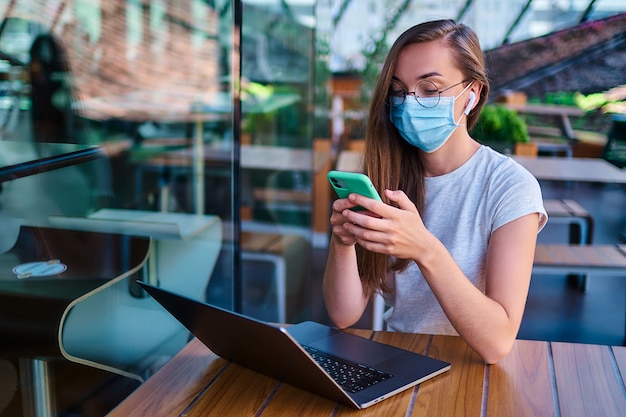 Libero professionista giovane donna che lavora in maschera facciale, cuffie senza fili bianche e occhiali rotondi utilizzando telefono e computer per lavorare in remoto online nella caffetteria