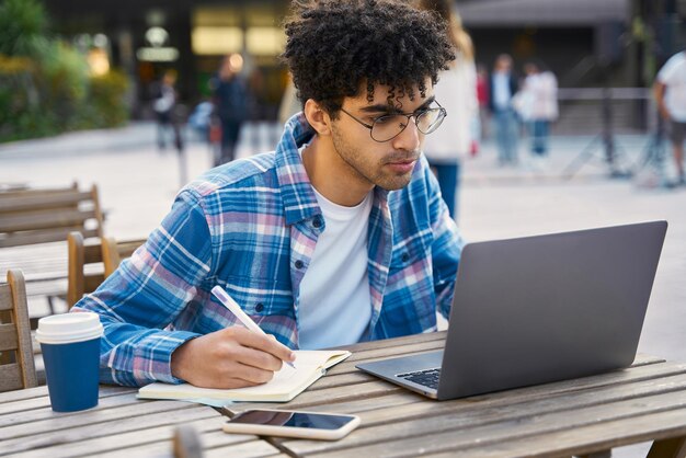 Libero professionista giovane uomo pensieroso utilizzando il computer portatile, prendendo appunti, guardando corsi di formazione