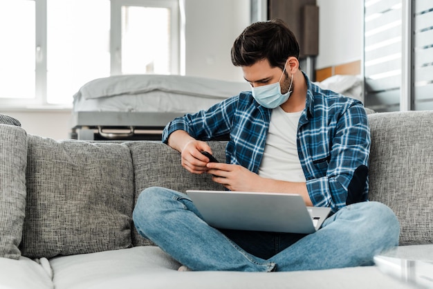 Libero professionista in maschera medica utilizzando smartphone e laptop sul divano di casa