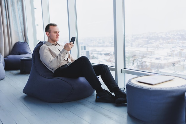 Libero professionista maschile di successo seduto in uno spazio di lavoro moderno e che controlla le notifiche sul telefono cellulare in abiti casual Lavoro a distanza