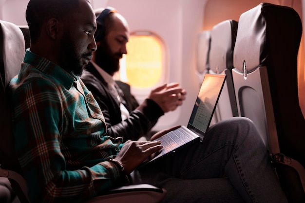 Libero professionista maschio afroamericano che lavora al computer portatile durante il volo al tramonto, in attesa di arrivare a destinazione di vacanza. Volare in classe economica con un gruppo di turisti, utilizzando il computer online.