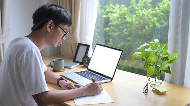 Libero professionista maschio asiatico concentrato che lavora online con il laptop e prende appunti sul taccuino