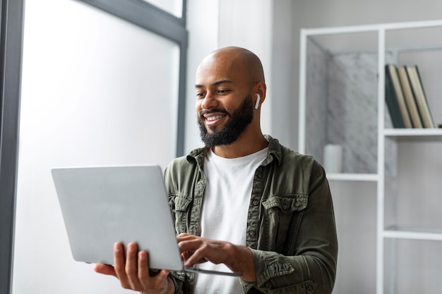 Libero professionista maschio felice che utilizza il computer portatile e indossa gli auricolari in piedi vicino alla finestra e digita sulla tastiera