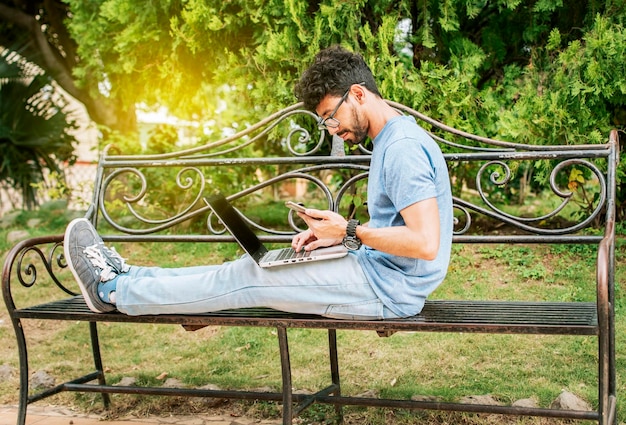 Libero professionista seduto in un parco con laptop e cellulare Uomo in un parco che lavora online con laptop Uomo rilassato che lavora con laptop all'aperto Giovane in un parco con laptop e telefono cellulare