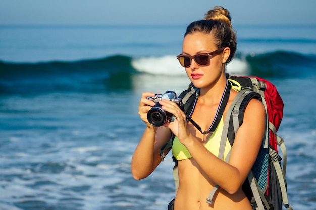 Libertà Giovane donna zaino in spalla in un costume da bagno bikini giallo sexy che tiene una macchina fotografica in mano su una spiaggia. donna bionda del fotografo in piedi vicino al mare con una grande borsa turistica sulle spalle, mare turistico