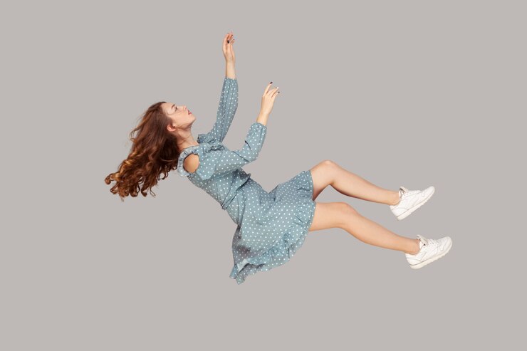 Librandosi nell'aria. Rilassata bella ragazza volant vestito e capelli ricci svettanti levitando, volando in sogno con le mani alzate, raggiungendo qualcosa di alto. indoor studio shot isolato su sfondo grigio