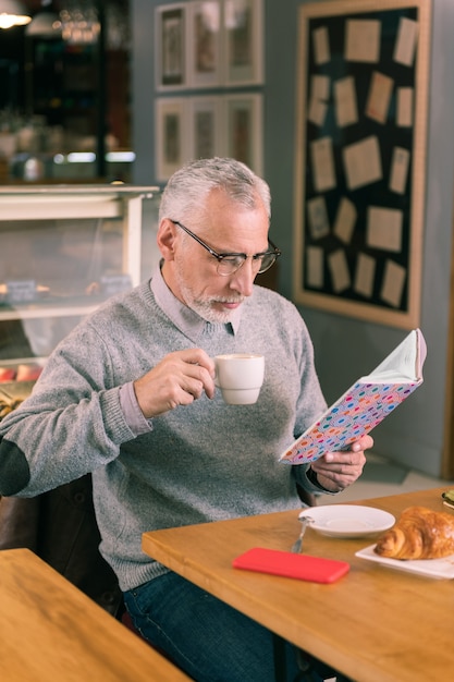 Libro da leggere. Barbuto uomo maturo con gli occhiali da lettura libro seduto in panetteria e bere caffè