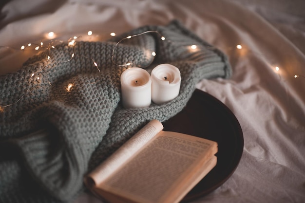 Libro di carta aperto su vassoio di legno con maglione lavorato a maglia con candele sopra luci incandescenti primo piano in camera. Orario serale invernale.