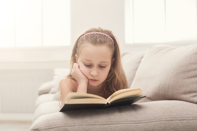 Libro di lettura carino bambina sdraiato sul divano di casa. Concetto di educazione e tempo libero, spazio di copia